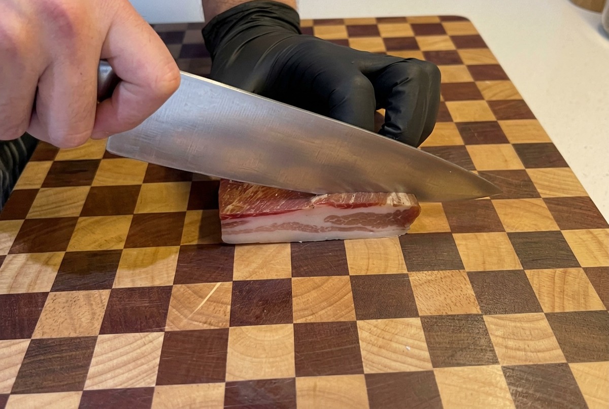 Hände mit schwarzen Handschuhen schneiden ein Stück Pancetta in kleine Würfel auf einem Holzschneidebrett.