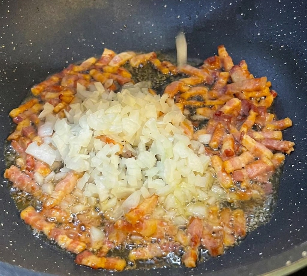 Fein gewürfelte weiße Zwiebeln werden in eine schwarze Pfanne zu der bratenden Pancetta gegeben.