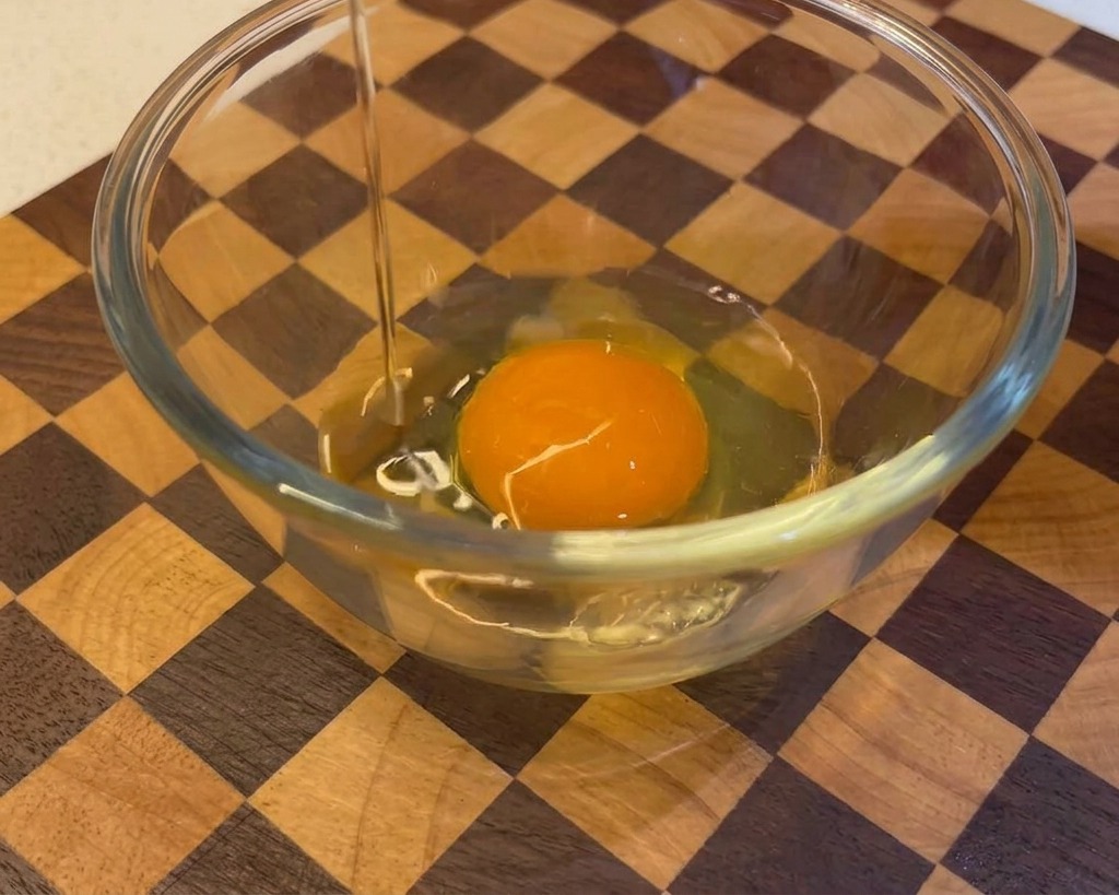 Ein rohes Ei wird aufgeschlagen und in eine Glasschüssel gegeben, in der sich bereits ein Eigelb befindet.