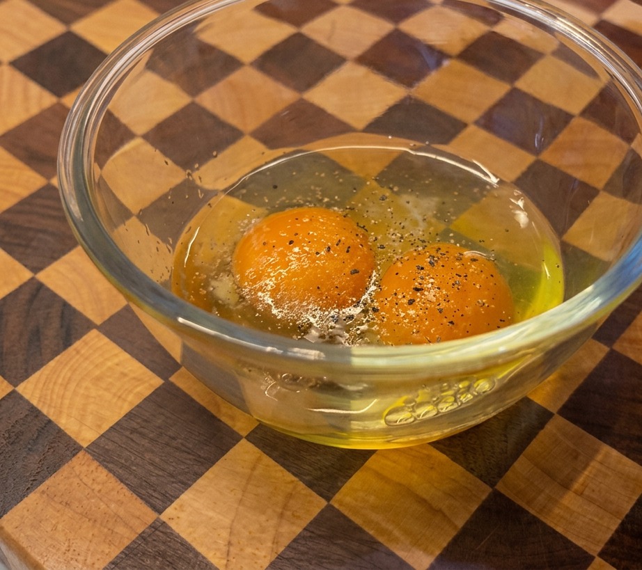 Eine Person mahlt schwarzen Pfeffer aus einer Holzmühle in eine Glasschüssel mit zwei orangefarbenen Eigelb.