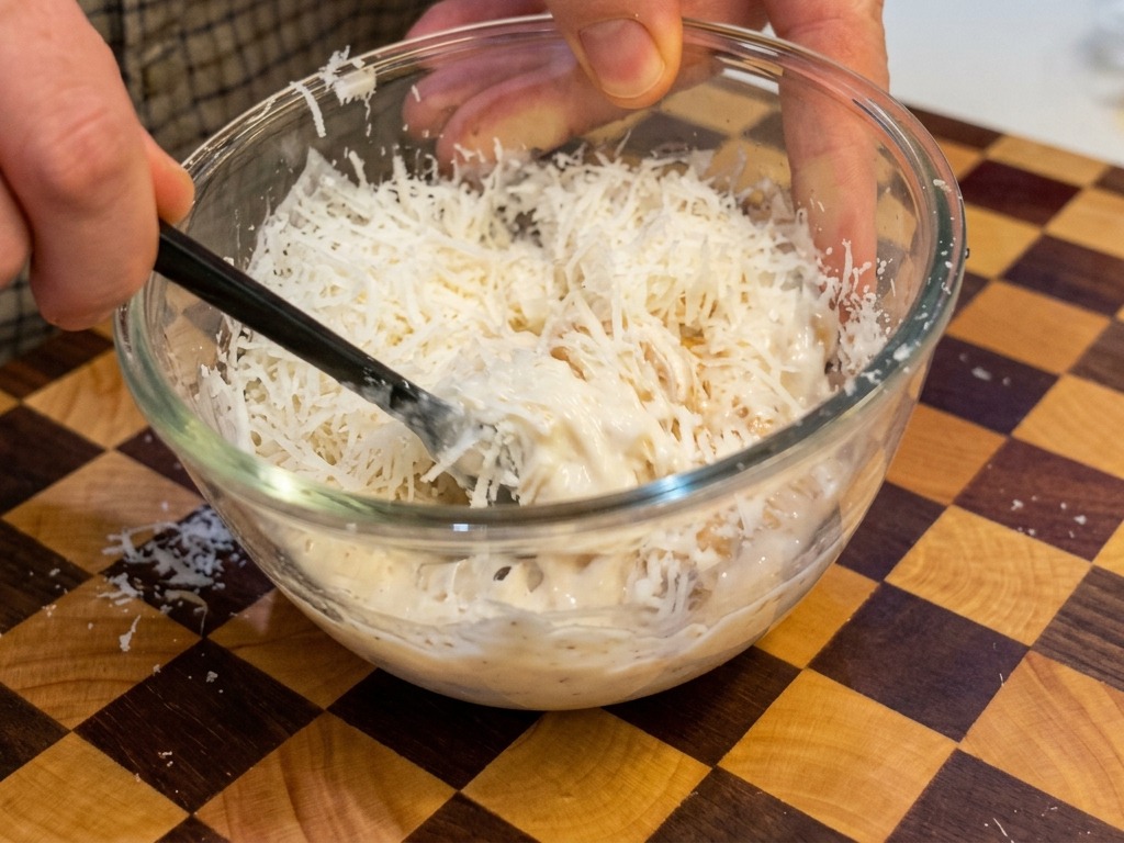 Eine dicke, cremige, blassgelbe Mischung aus Eiern und Käse wird mit schwarzen Stäbchen in einer klaren Schüssel verrührt.