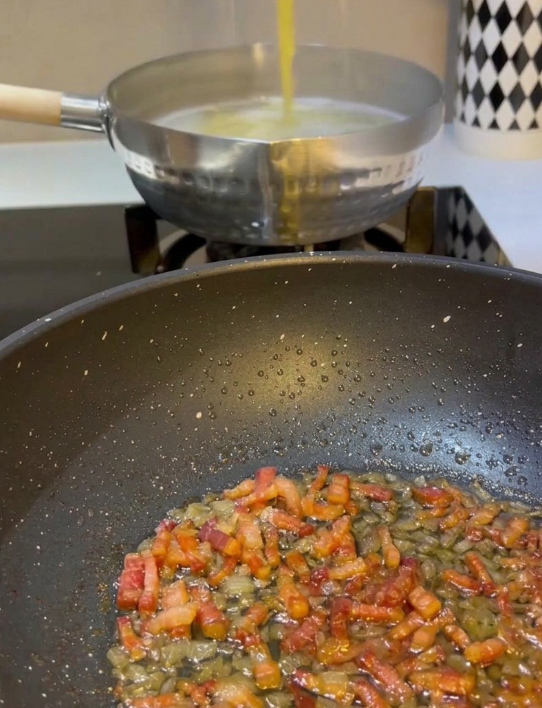 Eine Hand hebt mit einer Metallzange gelbe Spaghetti aus einem Topf mit kochendem Wasser direkt in eine Pfanne mit gebratener Pancetta und Zwiebeln.