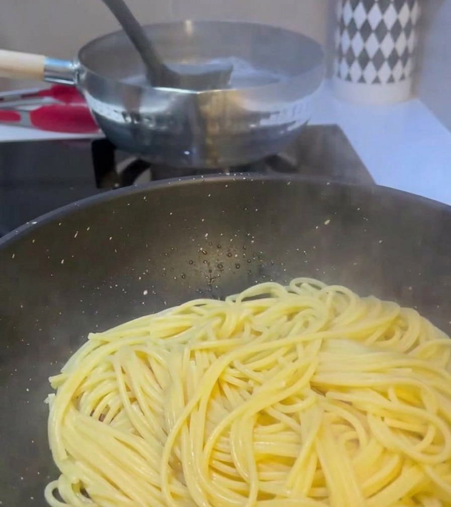Eine Kelle gießt trübes Nudelwasser aus einem kochenden Topf in eine Pfanne mit Spaghetti und gebratenem Fleisch.