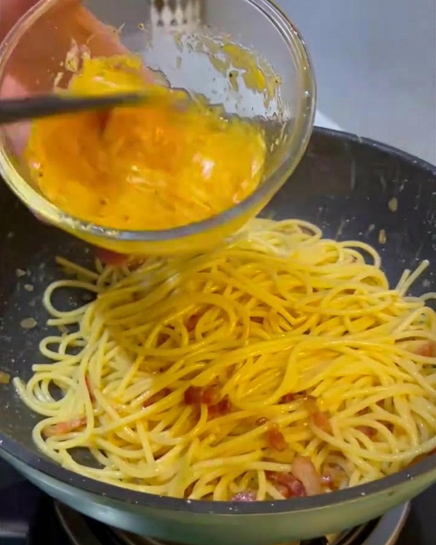 Eine kräftig gelbe Mischung aus Ei und Käse wird aus einer Glasschüssel in eine Pfanne mit heißen Spaghetti und gebratenem Fleisch gegossen.