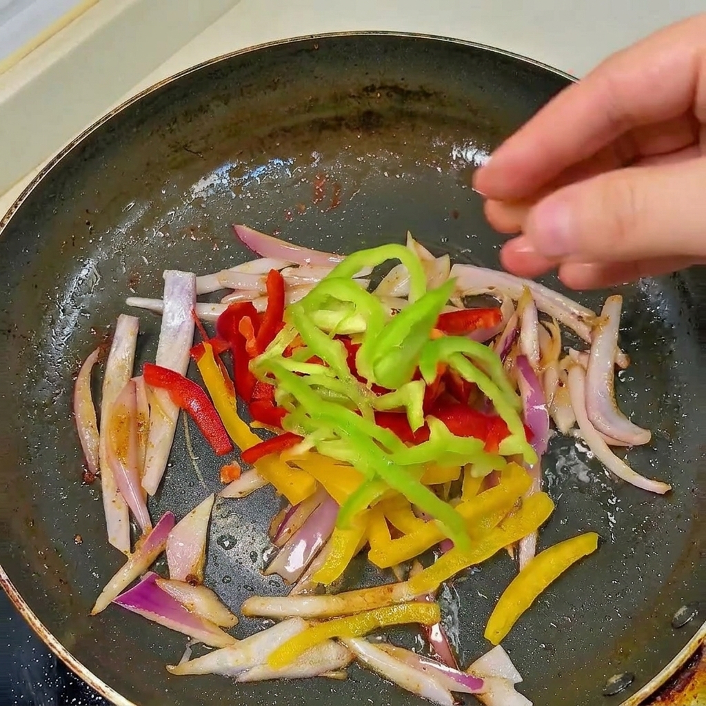 Eine Hand gibt frische rote, gelbe und grüne Paprikastreifen zu den angedünsteten Zwiebeln in die schwarze Pfanne.