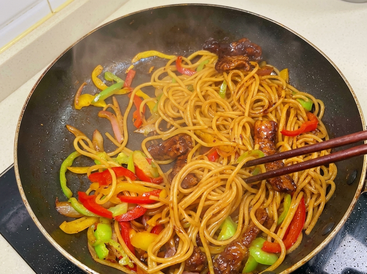 Mit Stäbchen wird die dampfende Mischung aus Spaghetti, Fleisch und Paprikastreifen in der glänzenden, dunklen Sauce angehoben.