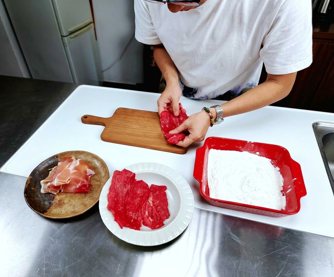 Dünne Scheiben von rohem, rotem Kalbfleisch flach ausgelegt auf einem hellen Holzschneidebrett auf einer Edelstahlarbeitsfläche.