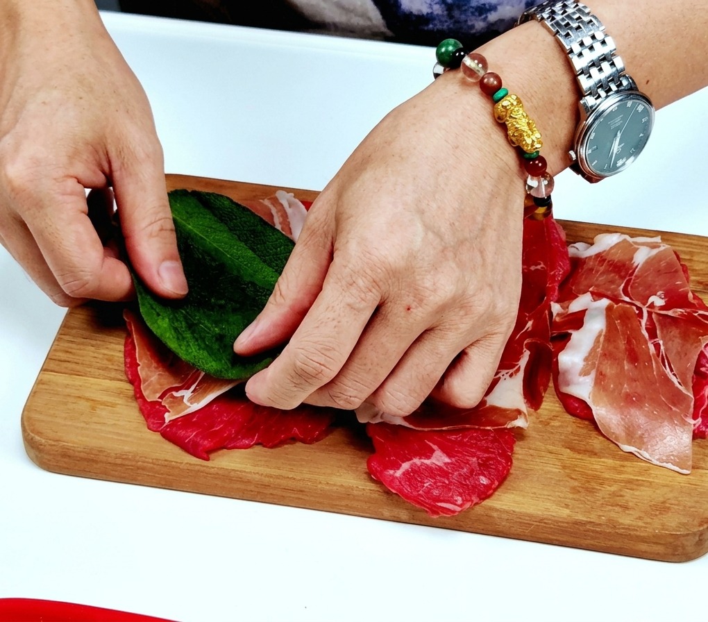 Hände, die ein großes, frisches grünes Salbeiblatt auf ein mit Prosciutto belegtes Kalbsschnitzel auf einem Holzbrett platzieren.