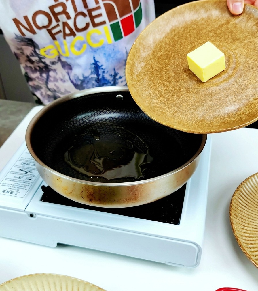 Ein Stück gelbe Butter wird in eine Edelstahlpfanne gegeben, in der sich bereits Olivenöl befindet.