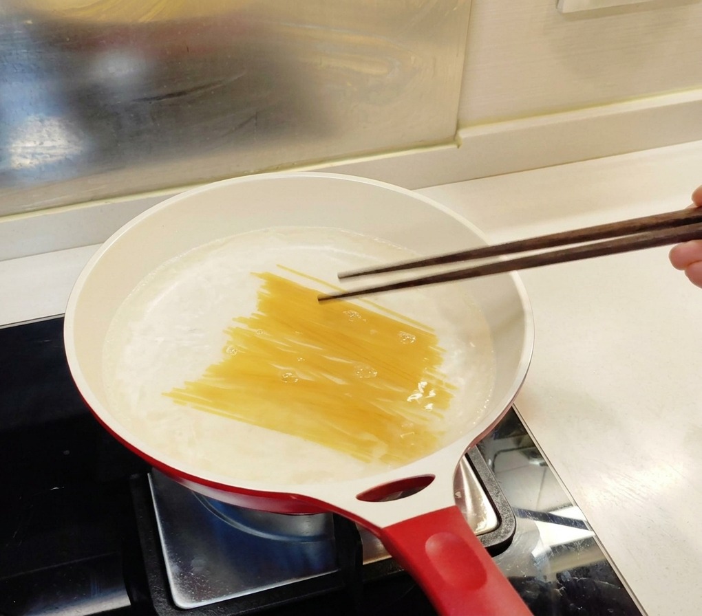 Trockene Spaghetti werden mit dunklen Holzstäbchen in einen weißen Topf mit kochendem Wasser gedrückt.