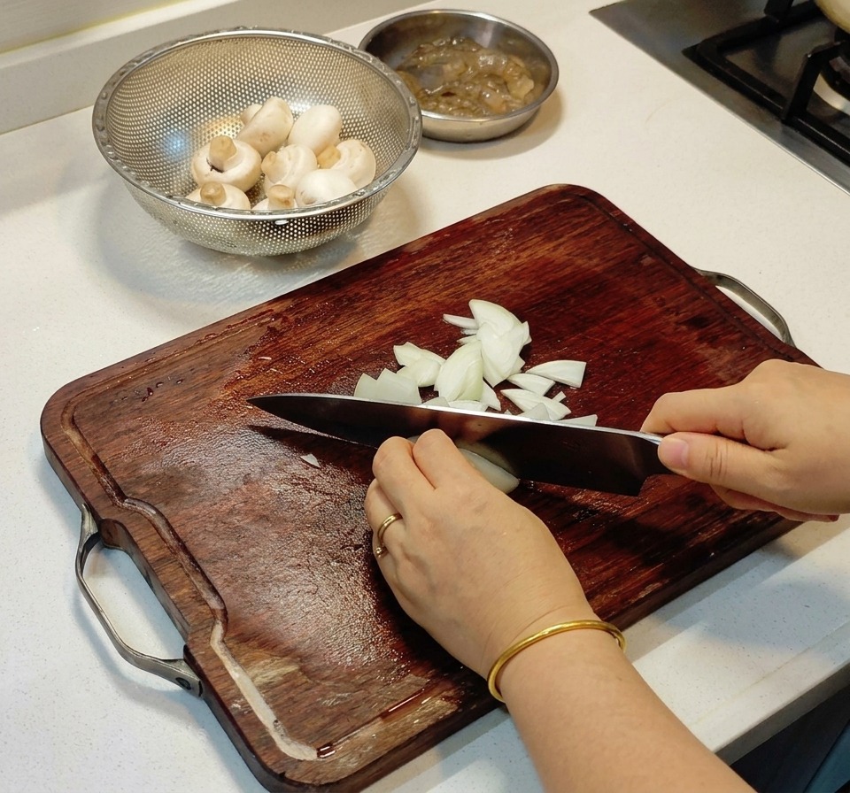 Hände würfeln eine weiße Zwiebel fein mit einem Kochmesser auf einem dunklen Holzschneidebrett neben einer Schale mit Pilzen.