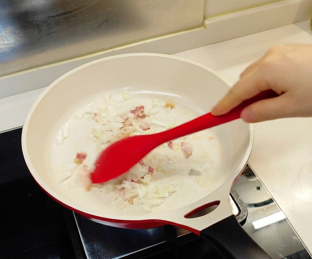 Ein roter Pfannenwender rührt gehackte Zwiebeln und kleine Speckstücke in einer weißen, keramikbeschichteten Pfanne.