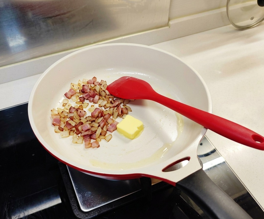 Ein einzelnes Stück gelbe Butter liegt in einer weißen Pfanne mit angebratenem Speck und Zwiebeln.