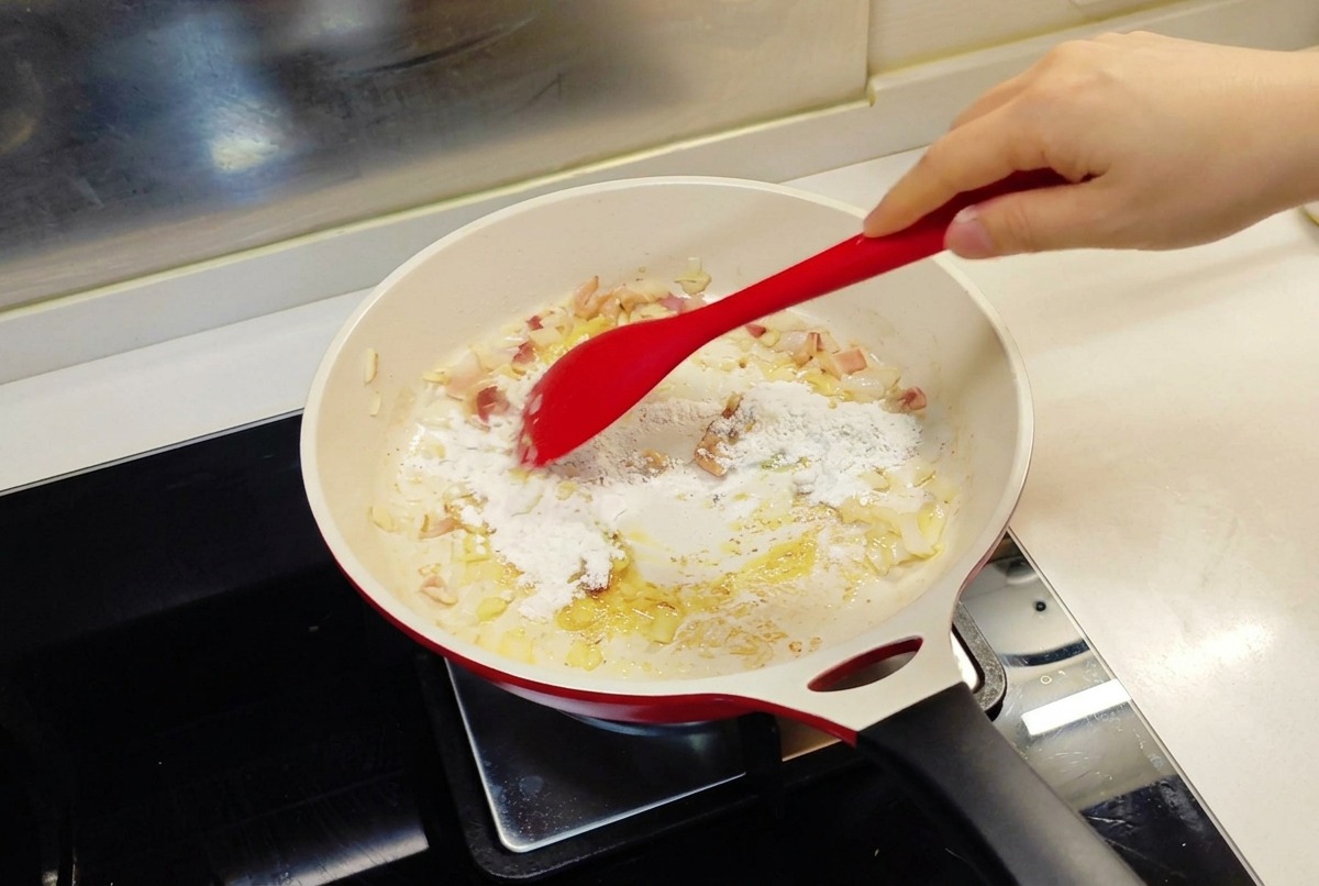 Ein roter Pfannenwender rührt weißes Mehl in eine Pfanne mit angebratenen Zwiebeln und geschmolzener Butter.