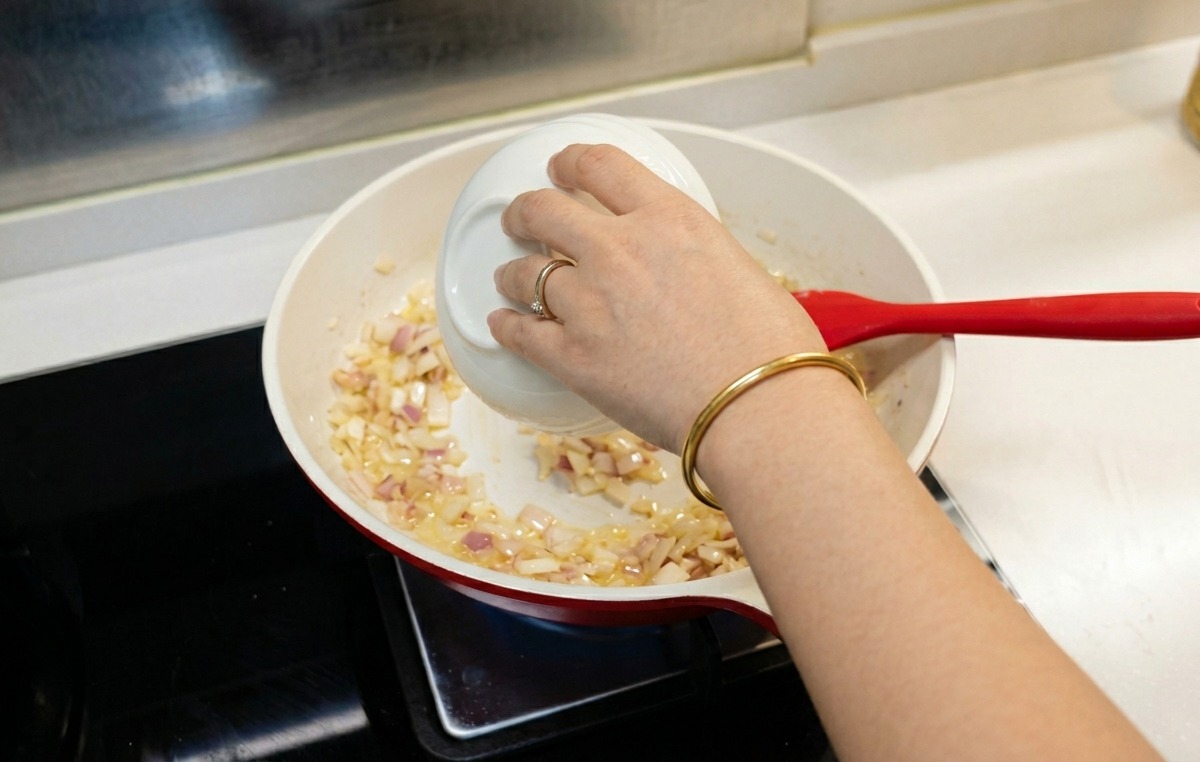 Eine Person gießt Wasser aus einer kleinen weißen Schale in eine Pfanne mit gegarten Zwiebeln und Mehl.