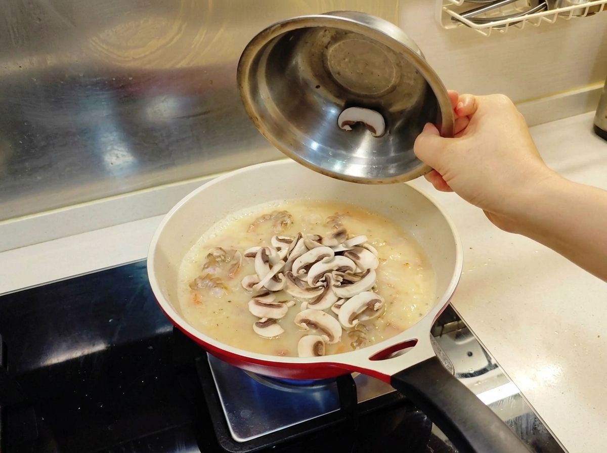 Frische weiße Champignonscheiben werden aus einer Metallschüssel in eine Bratpfanne mit einer cremigen Saucenbasis und Garnelen gegeben.