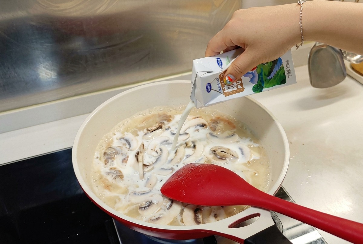 Milch wird aus einem weiß-blauen Karton in eine köchelnde Pfanne mit Garnelen und Pilzen gegossen.