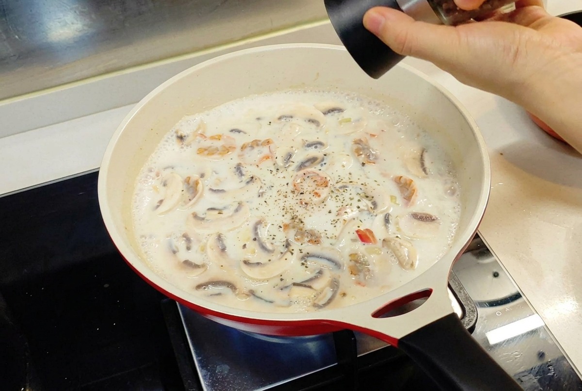 Eine Hand gibt mit einer Pfeffermühle schwarzen Pfeffer in eine cremige weiße Sauce, die in einer roten Pfanne köchelt.