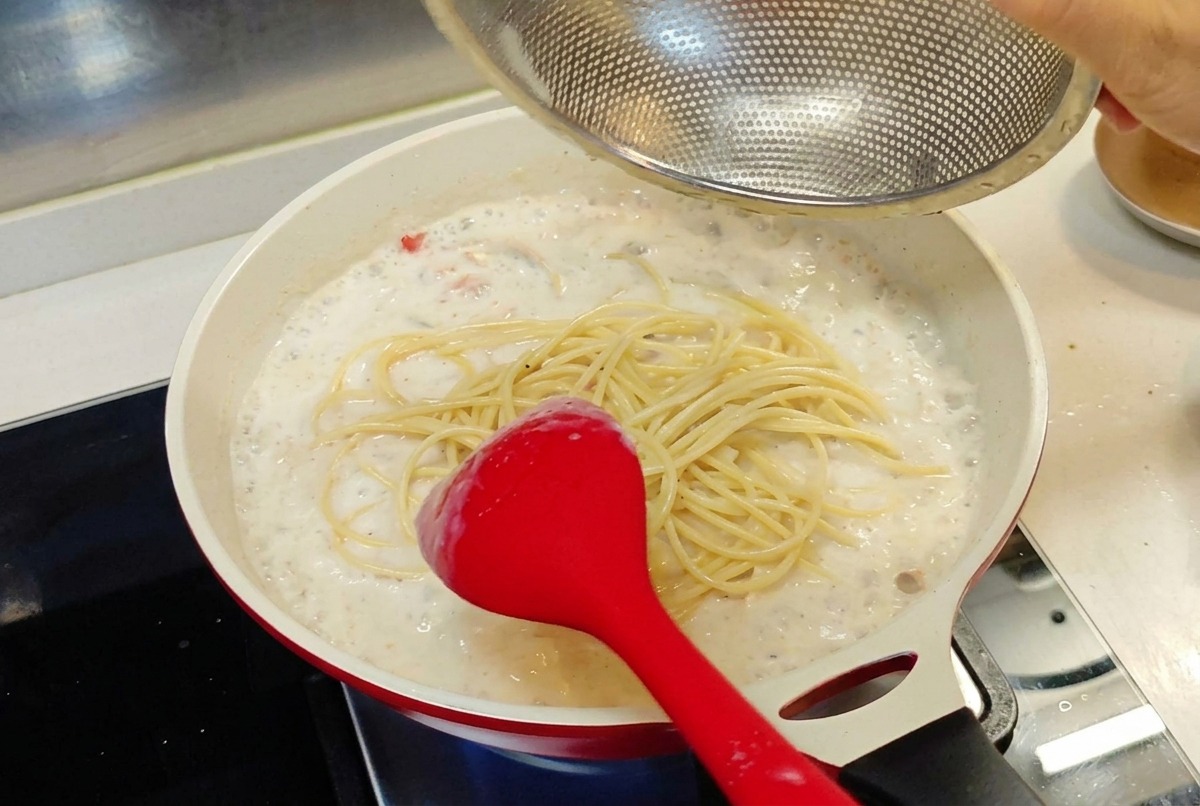 Gekochte Spaghetti werden aus einem Sieb in eine Pfanne mit cremiger weißer Sauce gegeben.
