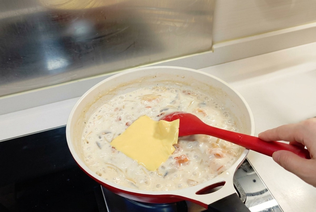 Eine quadratische Scheibe gelber Käse liegt auf einer brodelnden Pasta-Mischung mit weißer Sauce in einer weißen Pfanne mit einem roten Löffel.