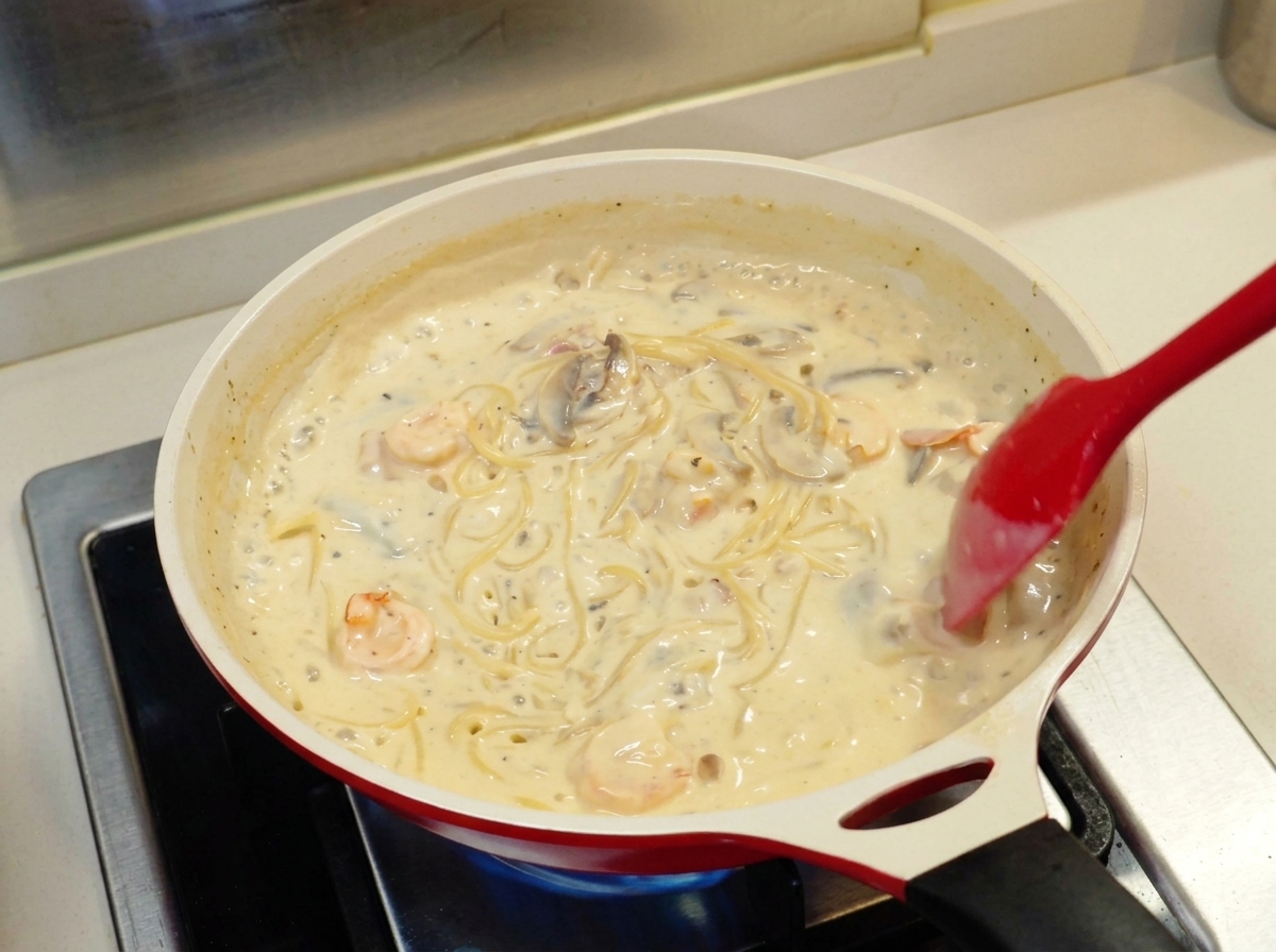 Ein roter Pfannenwender rührt Spaghetti in einer dicken, cremigen weißen Sauce mit Garnelen und Pilzen in einer weißen Keramikpfanne.