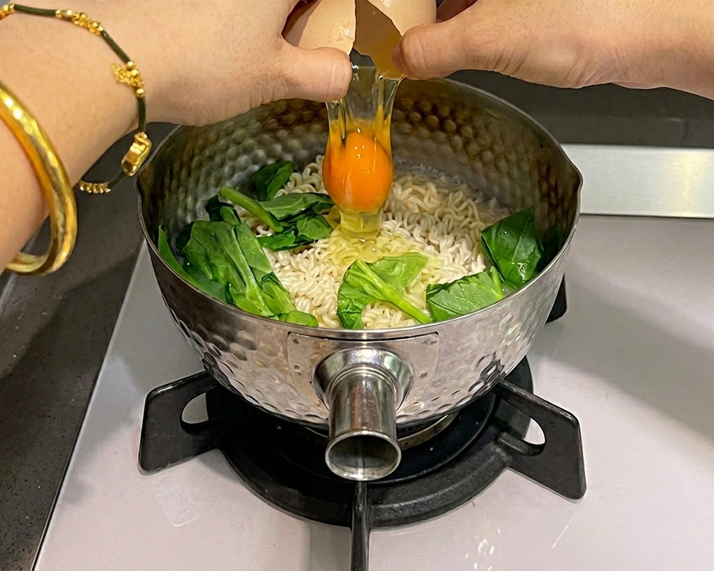 Zwei Hände schlagen ein rohes Ei über einem Topf mit kochenden Instant-Nudeln und grünem Spinat auf.