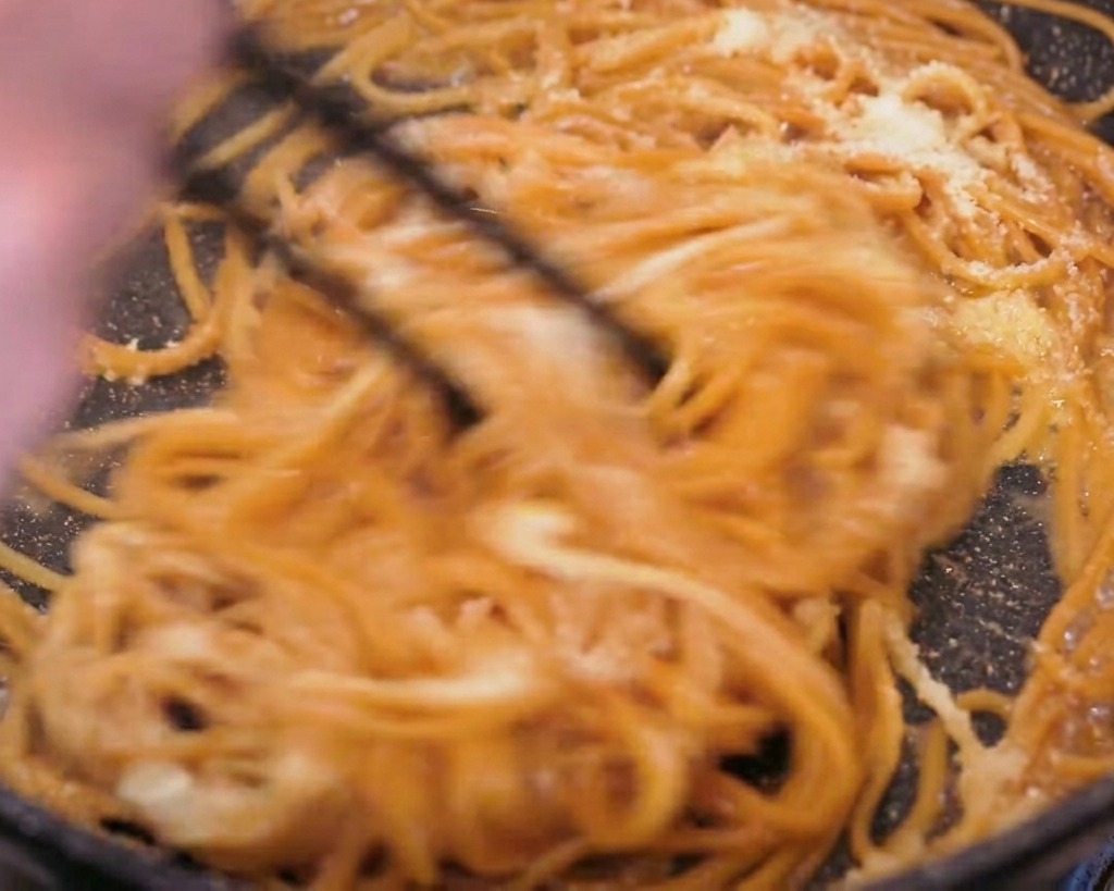 Action-Shot, bei dem Vollkornpasta mit schwarzen Stäbchen kräftig in der Pfanne geschwenkt wird, während Butter und Käse schmelzen.