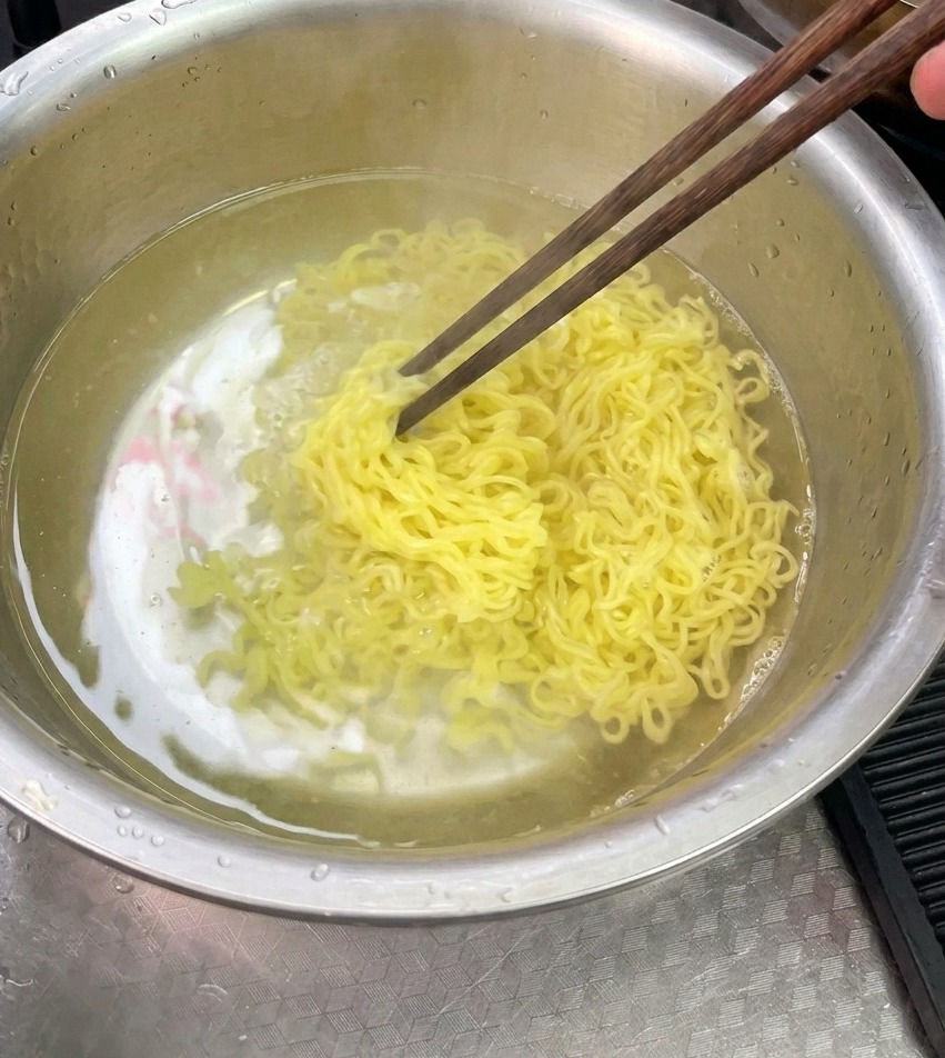 Gekochte gelbe Nudeln werden in einer Edelstahlschüssel mit kaltem Wasser abgespült und mit Essstäbchen umgerührt.