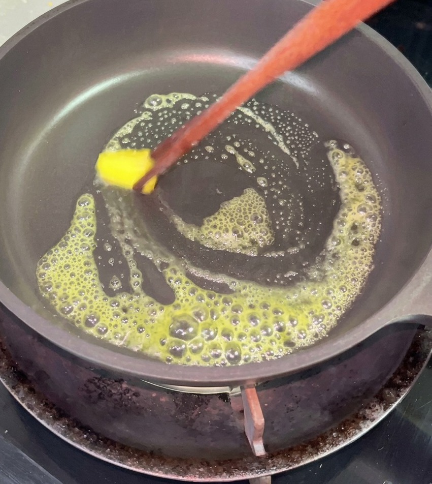 Ein kleines quadratisches Stück gelbe Butter schmilzt schäumend in einer schwarzen, beschichteten Pfanne und wird mit Essstäbchen bewegt.