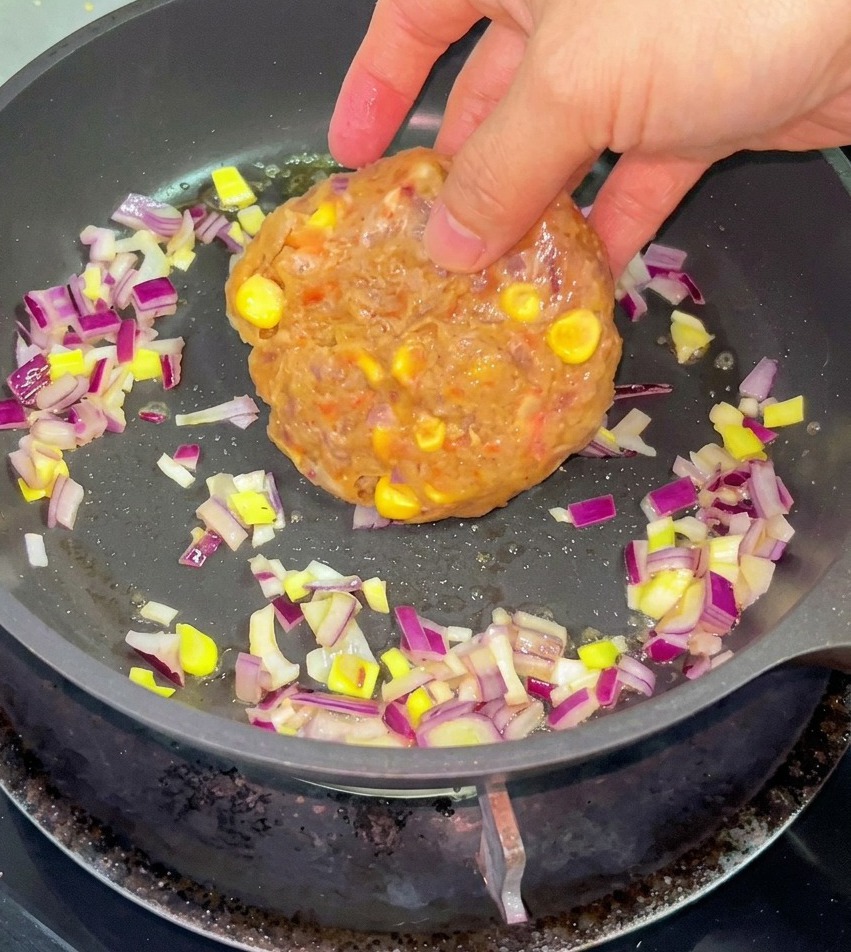 Eine Hand legt ein rohes Rindfleisch-Mais-Patty zusammen mit gehackten roten Zwiebeln in eine Pfanne mit geschmolzener Butter.