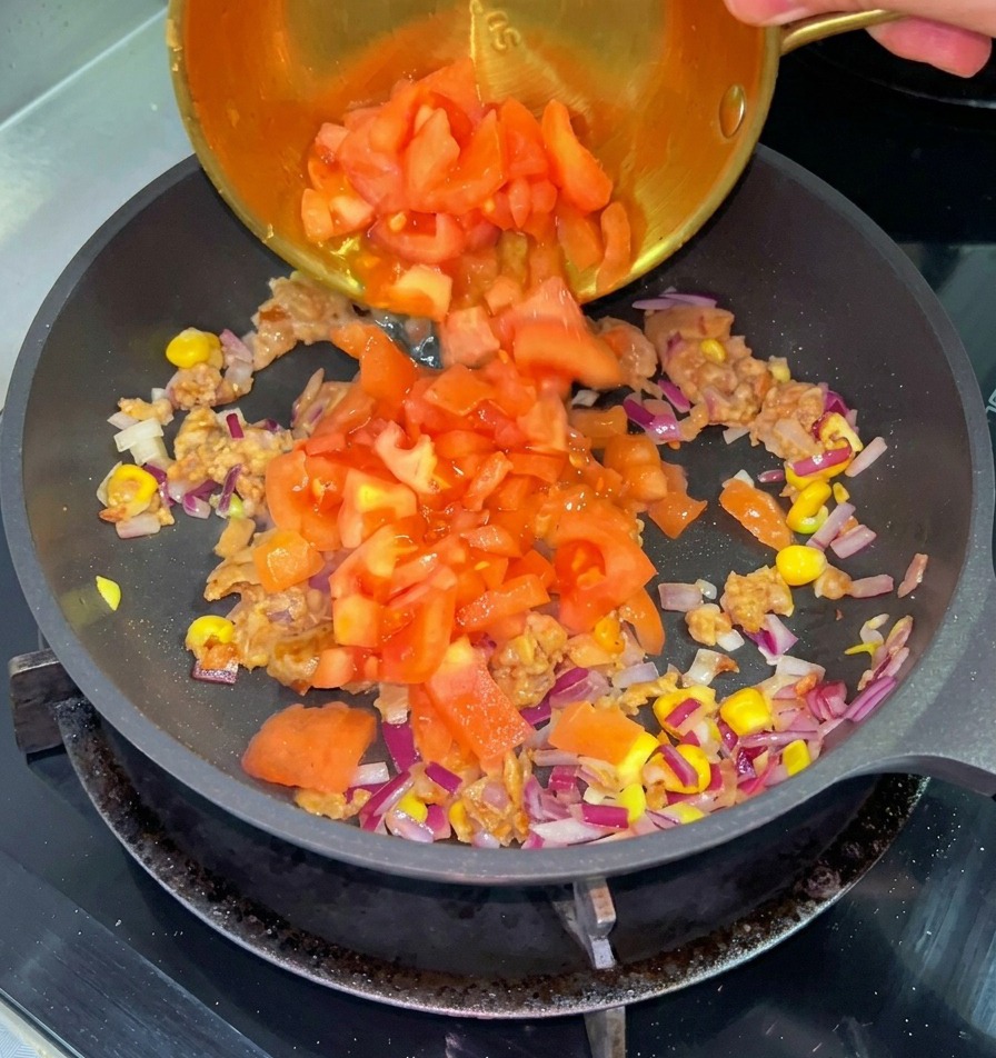 Gewürfelte rote Tomaten werden aus einer kleinen Messingschale in eine Pfanne mit angebratenem Rindfleisch, Mais und roten Zwiebeln geschüttet.