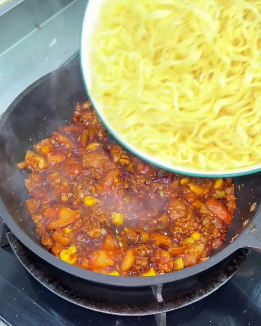 Eine Schüssel mit gewellten gelben Nudeln wird in eine Pfanne mit einer stückigen, dunkelroten Tomaten-Rindfleisch-Sauce geschüttet.
