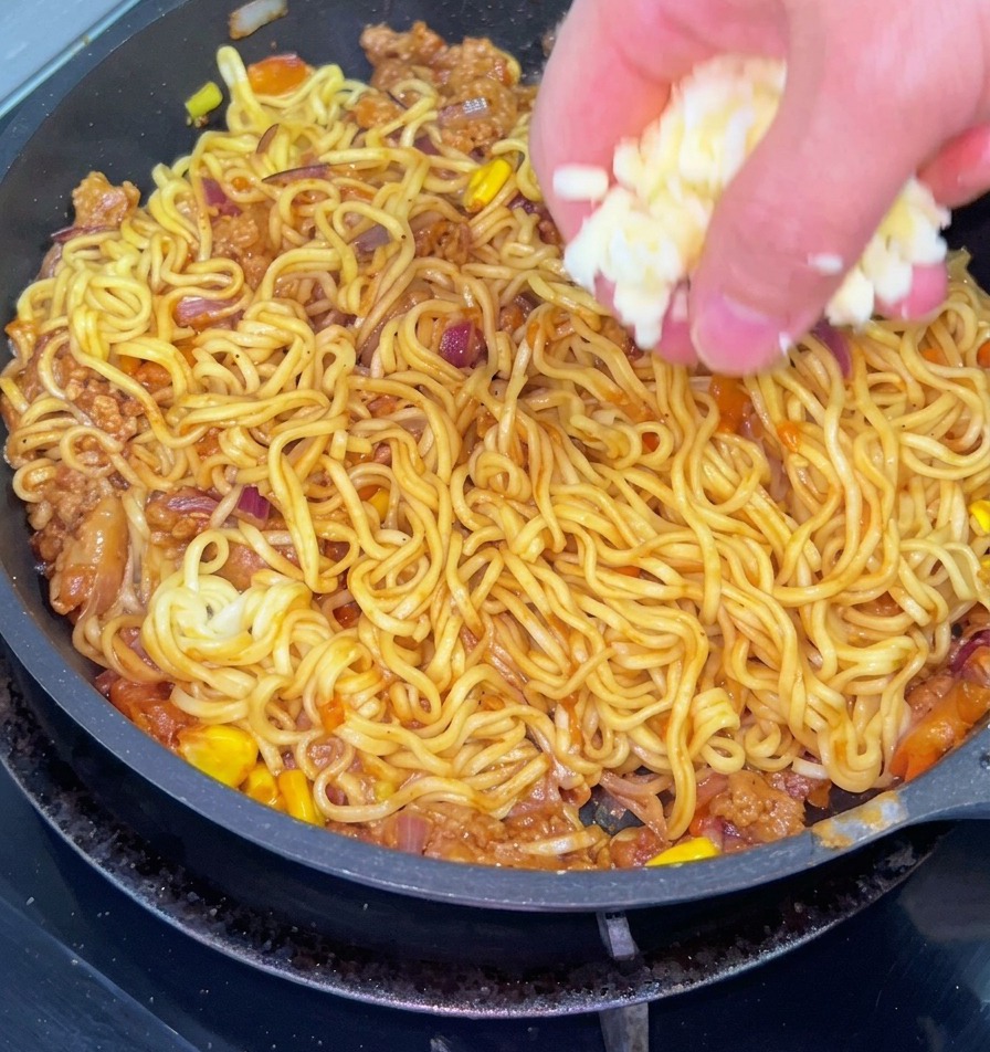 Eine Nahaufnahme zeigt eine Hand, die geriebenen weißen Käse über eine Pfanne mit gebratenen Nudeln streut, die mit Rinderhack, Zwiebeln und Mais vermischt sind.