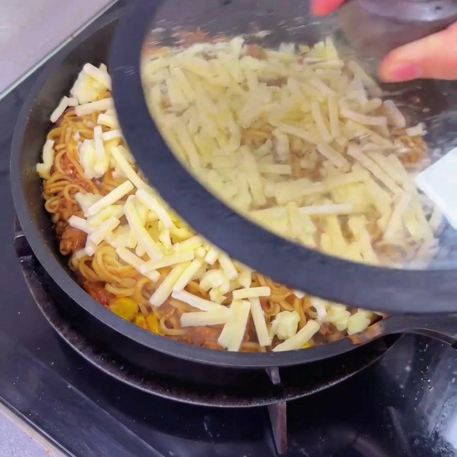 Eine Person setzt einen klaren Glasdeckel auf eine große Pfanne mit Rindfleisch-Nudeln, die mit einer Schicht noch ungeschmolzenem geriebenem Käse bedeckt sind.