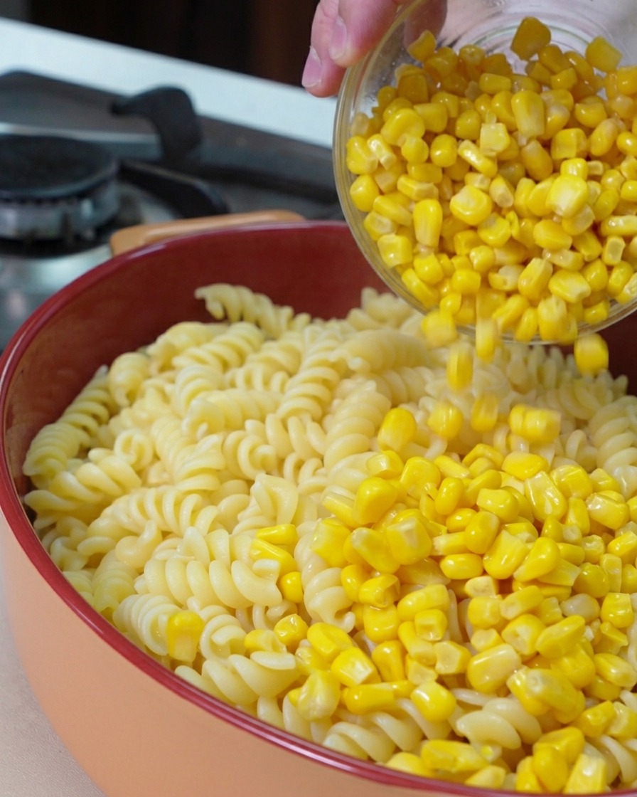 Aus einer Glasschale werden leuchtend gelbe Maiskörner über die gekochten Fusilli-Nudeln in einer runden Auflaufform gestreut.