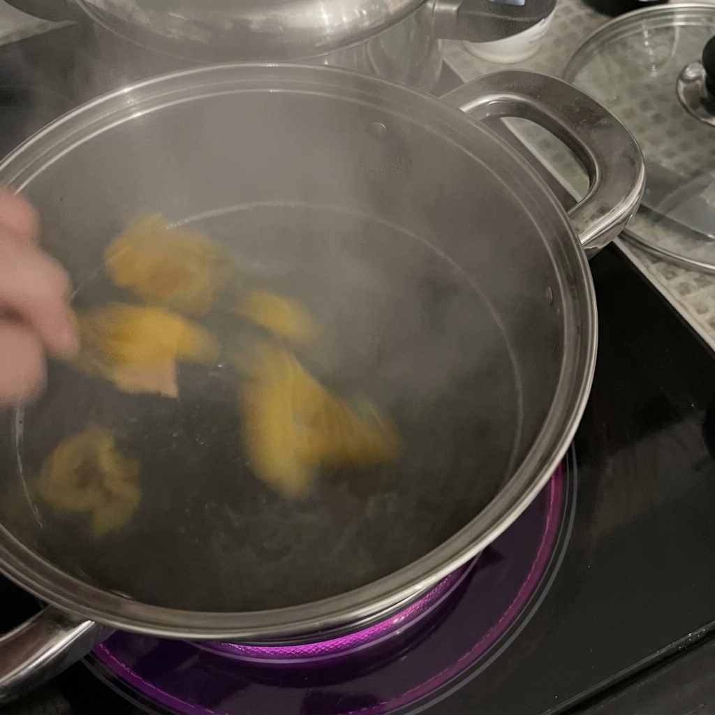 Eine Hand gibt gelbe Rigatoni-Nudeln in einen Edelstahltopf mit kochendem Wasser auf einem Induktionskochfeld.