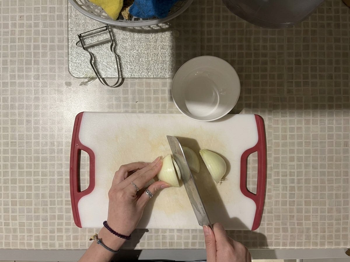 Draufsicht auf Hände, die eine weiße Zwiebel mit einem großen Messer auf einem weißen Schneidebrett mit roten Griffen in Würfel schneiden.