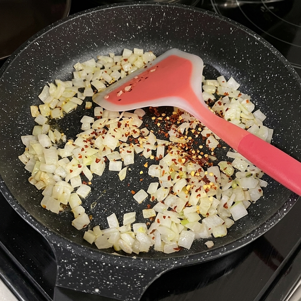 Rote getrocknete Chiliflocken sind über glasig gedünsteten Zwiebelwürfeln in einer schwarzen Pfanne verstreut.