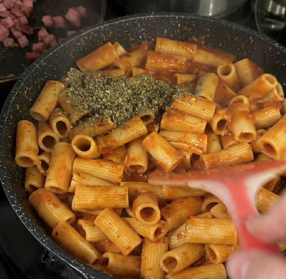 Ein großer Haufen getrocknete grüne Basilikumflocken liegt auf den Rigatoni-Nudeln in Tomatensoße in einer schwarzen Bratpfanne.
