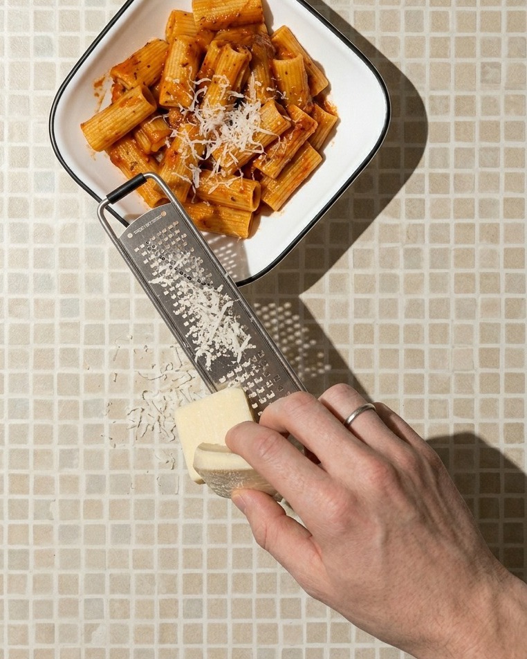 Nahaufnahme einer Hand, die ein Stück weißen Käse über eine weiße, quadratische Schüssel mit Rigatoni-Pasta in roter Soße reibt.