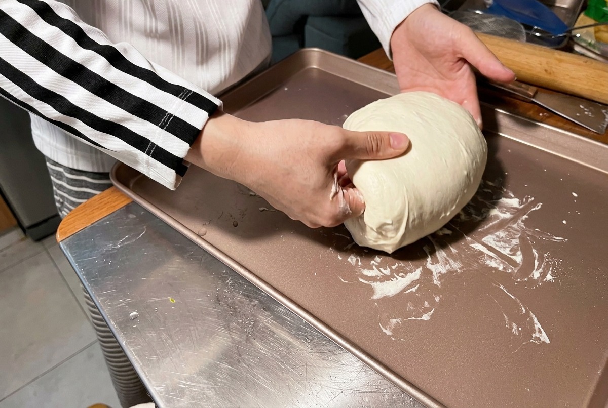 Eine Person formt mit den Händen eine große, weiche, weiße Teigkugel auf einem bronzefarbenen Backblech.