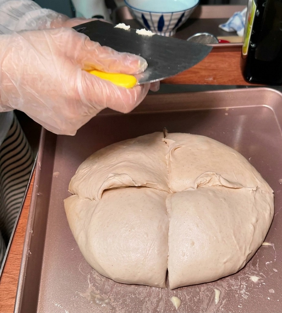 Eine Person mit einer Kunststoff-Handschuh teilt den großen Teig mit einem Teigschaber in kleinere, runde Portionen auf einem Blech auf.