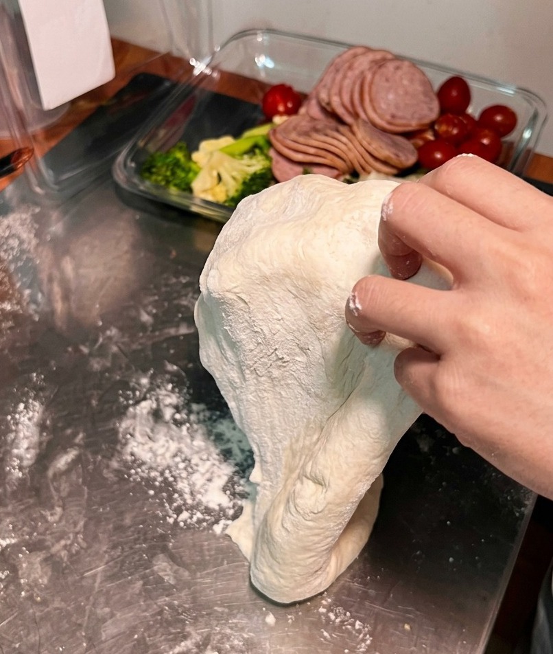 Ein Pizzateig wird über die Handrücken zweier Hände gelegt und gedehnt, um den Durchmesser zu vergrößern.