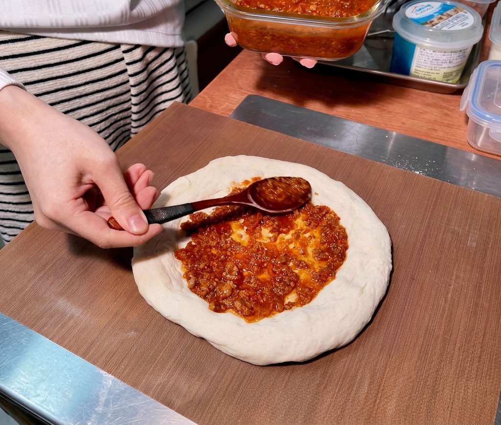 Ein Kochlöffel verteilt dicke, rötliche Hackfleischsauce in kreisenden Bewegungen auf einem rohen Pizzateigboden.