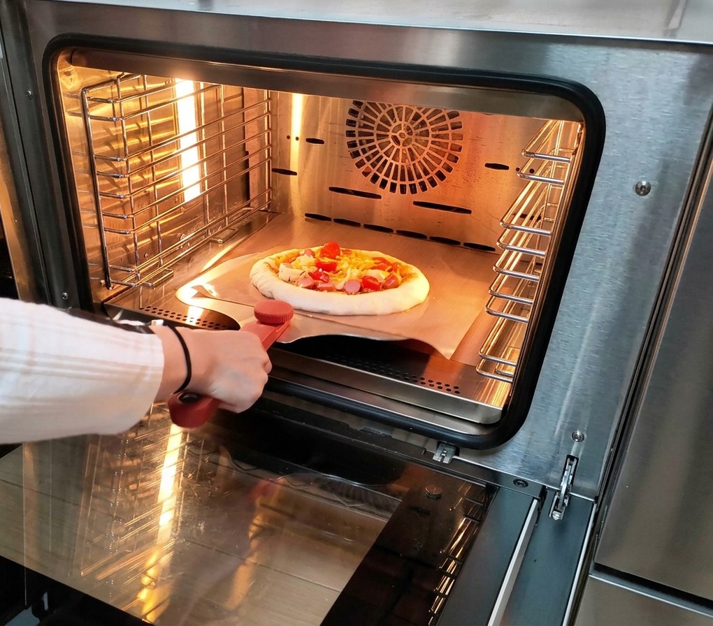 Eine Person schiebt eine rohe, reich belegte Pizza mithilfe eines Pizzaschiebers aus Metall in einen heißen, glänzenden elektrischen Ofen.