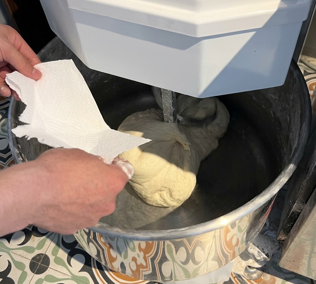 Eine Hand gibt Salz aus einer Papiertüte in eine Rührschüssel mit einer großen, glatten Pizzateigkugel.
