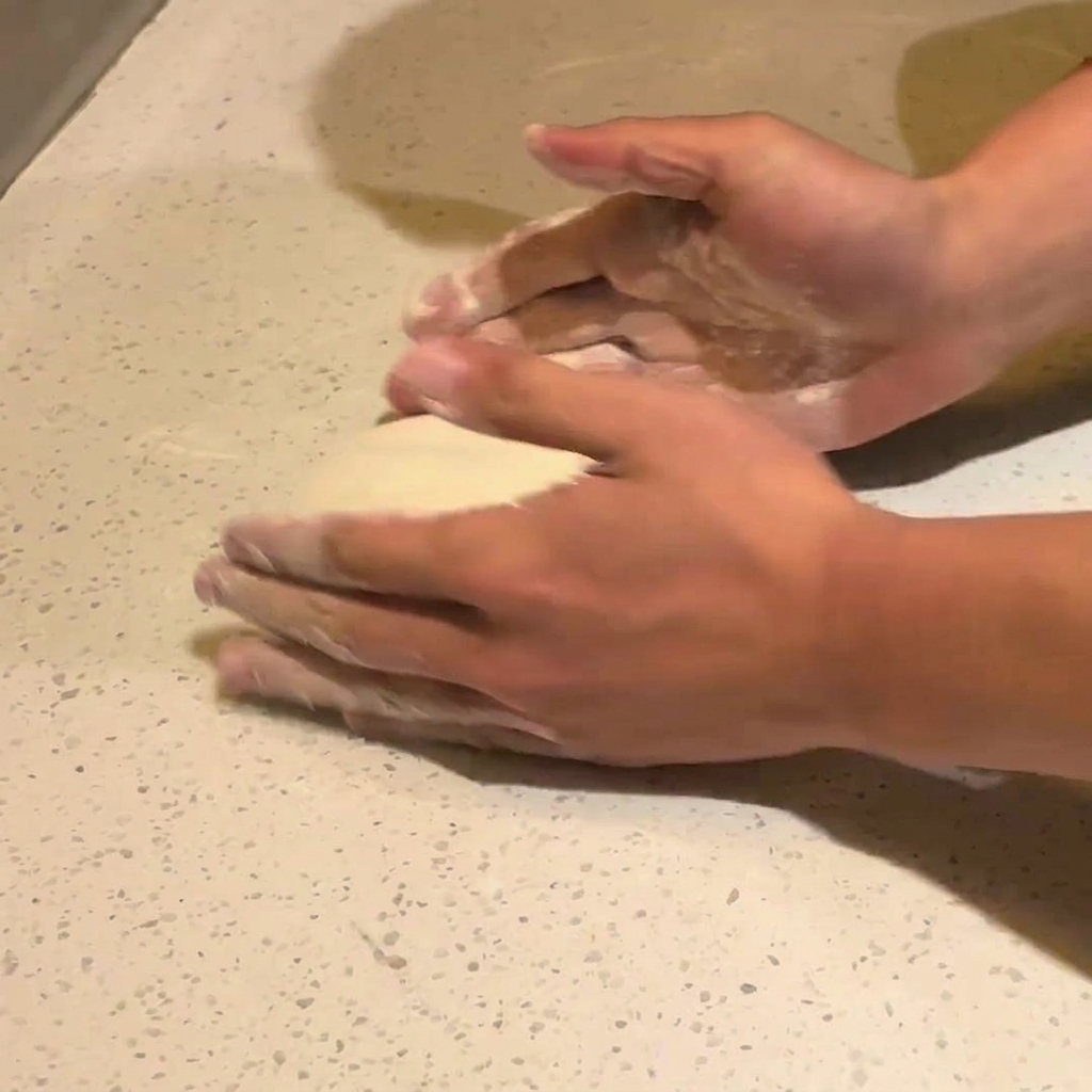 Zwei Hände drücken auf die Mitte eines runden Stücks fermentierten Pizzateigs auf einer bemehlten weißen Arbeitsfläche.