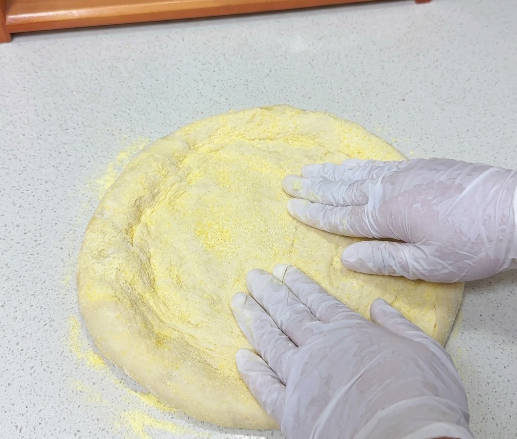 Hände drücken auf ein rundes Stück Pizzateig, das mit gelbem Grieß auf einer hellen Arbeitsfläche bedeckt ist.