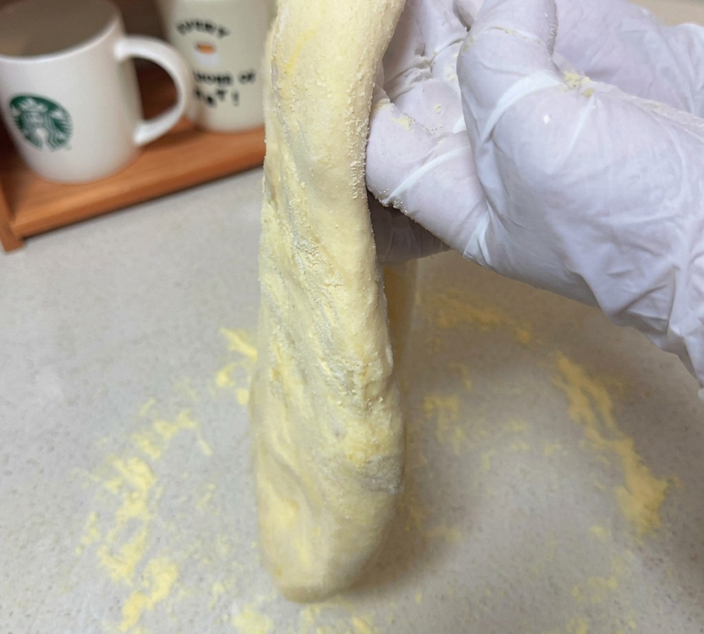 Ein langgezogenes Stück roher Pizzateig wird von Händen über einer bemehlten Fläche gehalten.