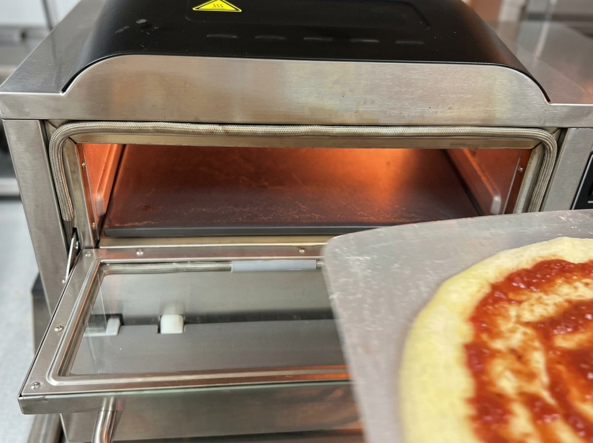 Eine rohe Pizza mit Tomatensauce und Mozzarella wird von einem Metall-Pizzaschieber in das glühende Innere eines elektrischen Pizzaofens geschoben.
